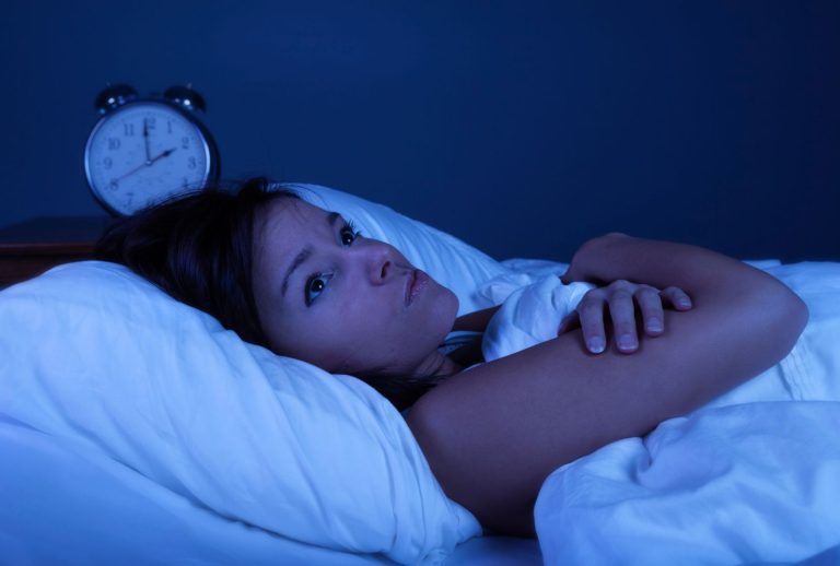 Mulher com insônia / dificuldade de dormir deitada na cama próxima a um despertador